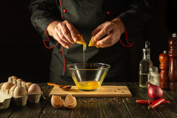 The Chef Pours A Raw Egg Into A Bowl. Delicious Breakfast Menu Idea For Hotel Or Restaurant On Dark Background